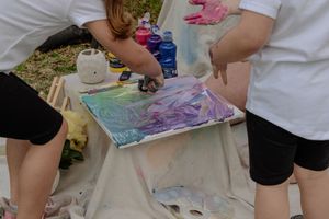 Children sit outdoors on a white sheet creating colorful art projects with various art supplies and materials.