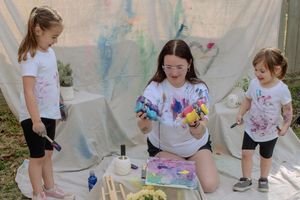 Children participate in a colorful outdoor art activity with paint and paper under a white tent canopy.