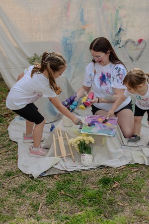 Three people sitting on a white cloth outdoors doing tie-dye art activities together on a sunny day.