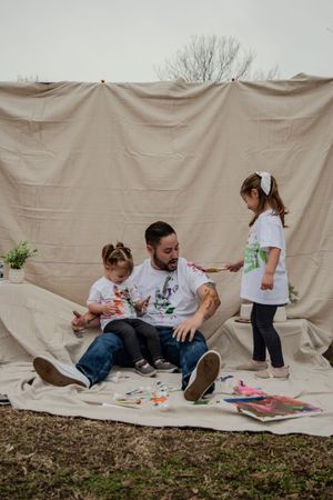 A family sits on a white backdrop outdoors doing a creative art activity together on the ground.