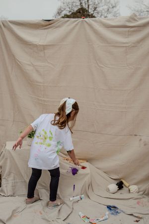 Young child in white t-shirt and leggings paints on large beige backdrop while creating messy art project outdoors.