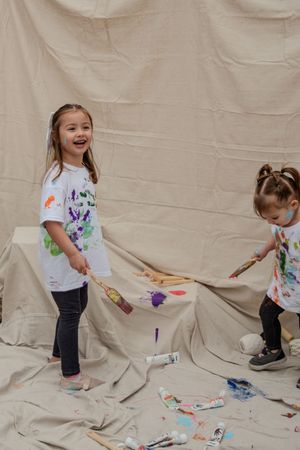 Child in white shirt and jeans plays with colorful paint on a beige backdrop creating artistic expression.