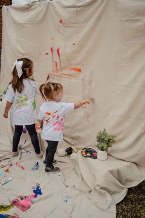 Children paint and draw on a large white canvas backdrop set up against a brick wall outdoors.