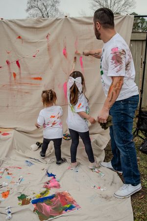 Family wearing matching white t-shirts painting together on a canvas backdrop in their backyard.