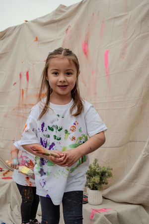 Young child wearing a paint-splattered white t-shirt smiles during a messy art activity against beige background.