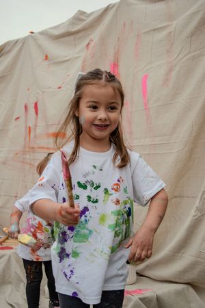 Young child in paint-splattered white t-shirt smiles while doing art activity against beige backdrop.