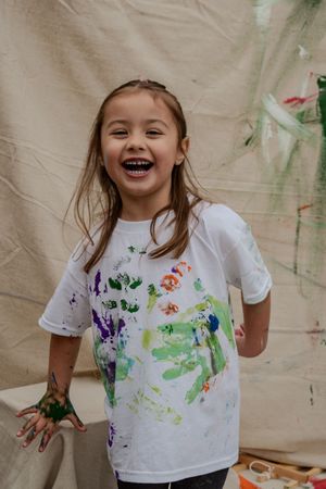 A child wearing a white t-shirt shows their hands and clothing covered in colorful paint splatters during an art activity.