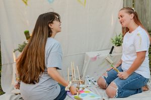 Two people sit together at a white table with colorful party decorations and a birthday cake in front of them.