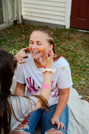 Friends having fun with colorful face paint and handprints on a white t-shirt during an outdoor gathering.