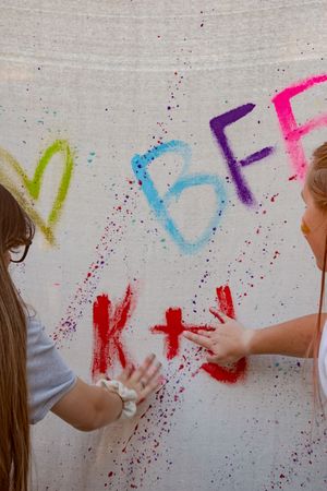 Colorful spray paint letters and splatters decorate a white backdrop as hands reach to create artistic graffiti.