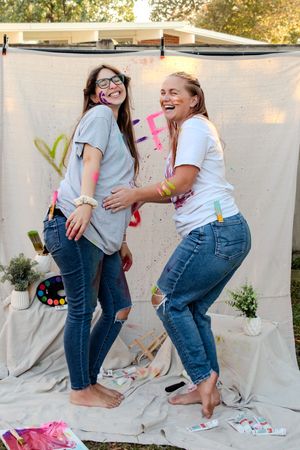Two friends in matching outfits share joyful moments while creating colorful art on a white backdrop outdoors.
