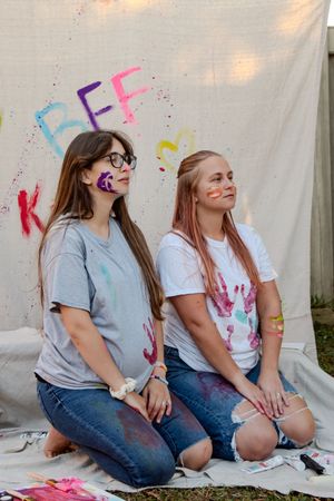 Two friends sit together on the ground with paint splatters on their clothes and wall during a colorful art session.
