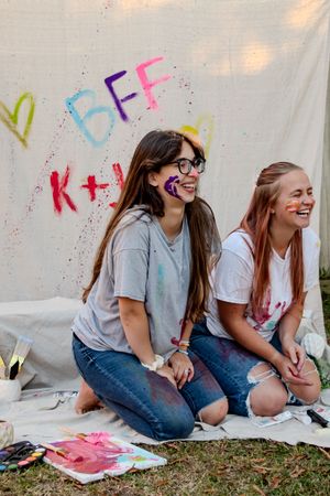 Friends in denim and t-shirts laugh together while sitting on the ground with colorful art materials.