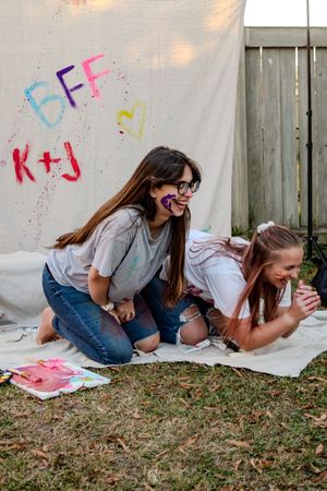 Two friends laughing together on a blanket with colorful spray paint art on a backdrop behind them.
