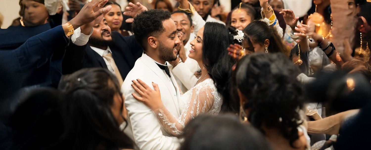 Couple sharing romantic first dance embrace surrounded by cheering wedding guests indoors.