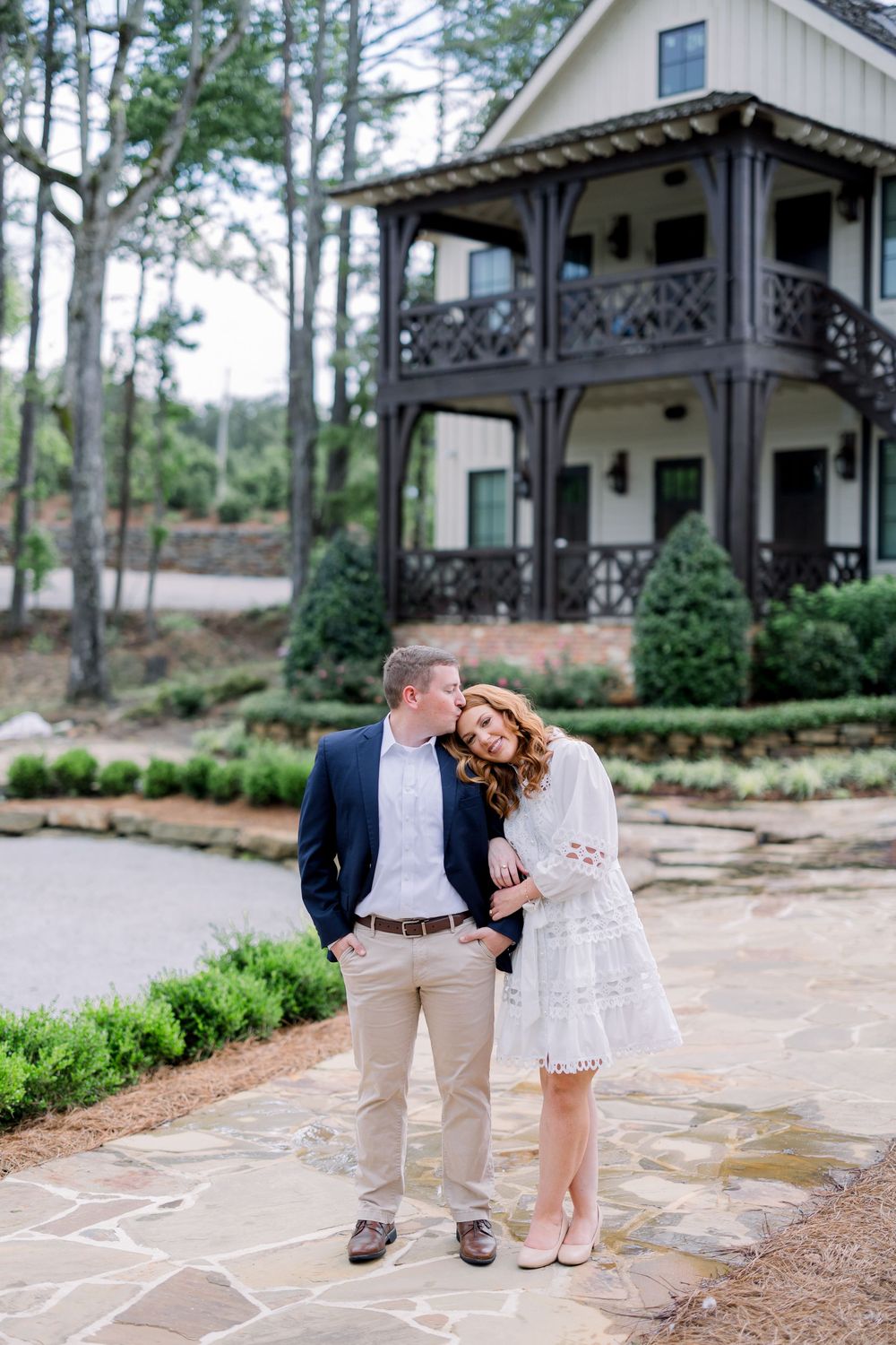 A couple strolls together on a flagstone walkway in front of a white farmhouse-style building with pine trees.
