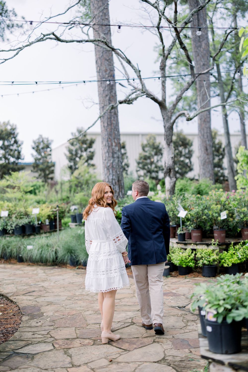 A couple shares a romantic moment on a stone patio surrounded by potted plants and tall pine trees in a garden setting.