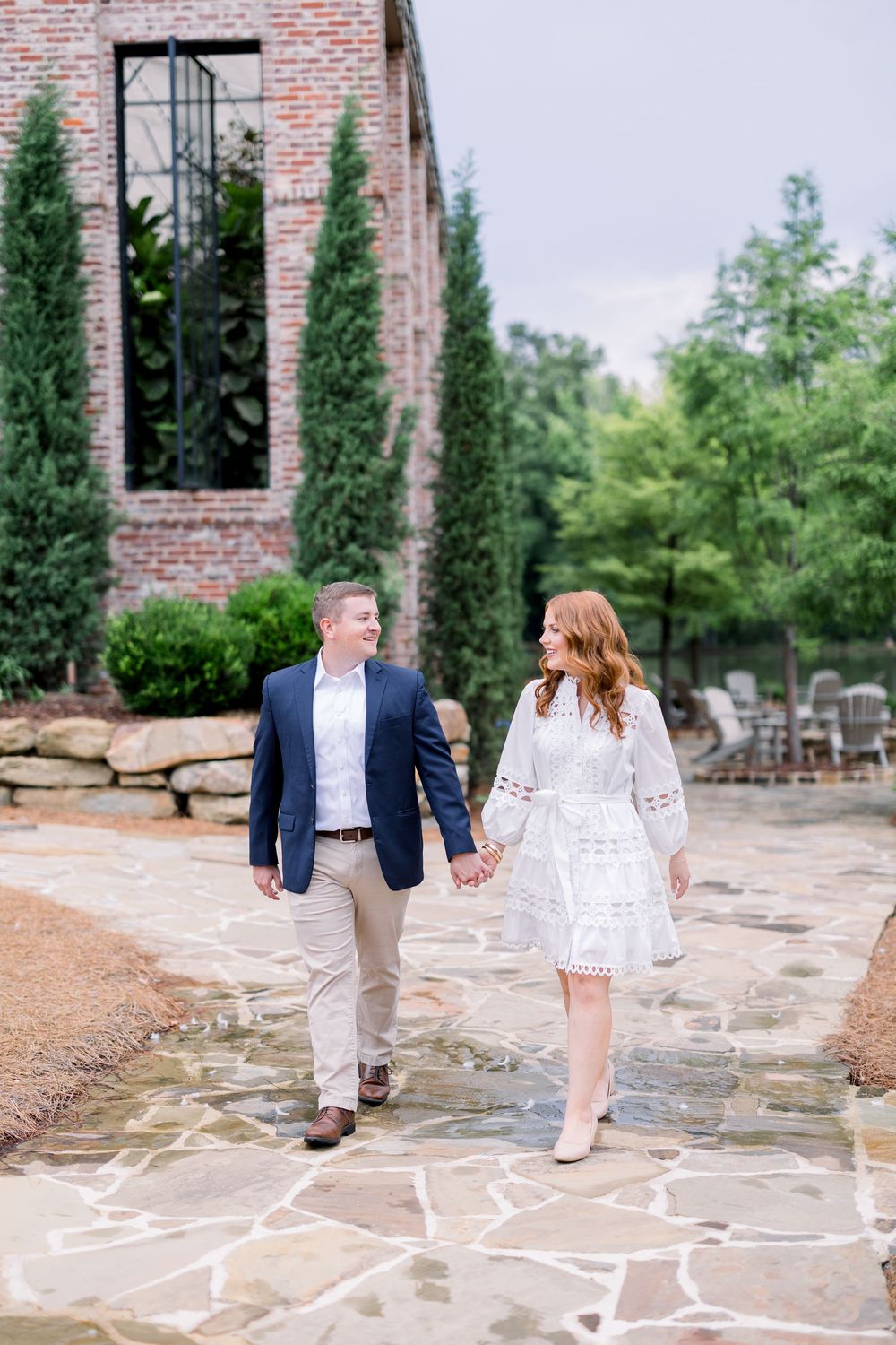A couple walks hand in hand on a stone patio path beside a brick building with tall cypress trees.
