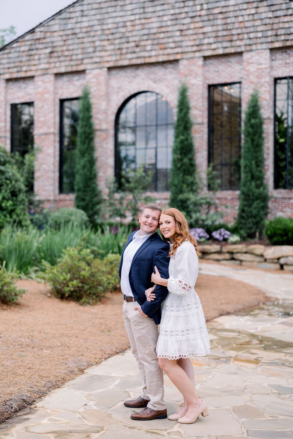 A romantic outdoor engagement photo session against a rustic brick building with landscaped greenery.