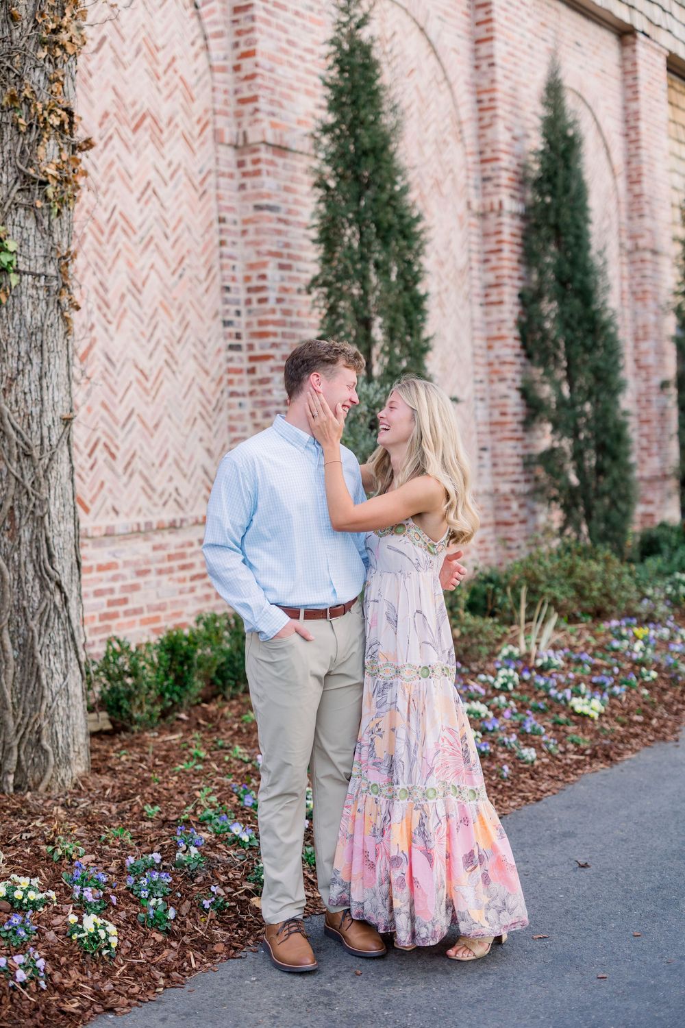 A couple shares romantic moments along a brick wall with cypress trees in an outdoor garden setting.