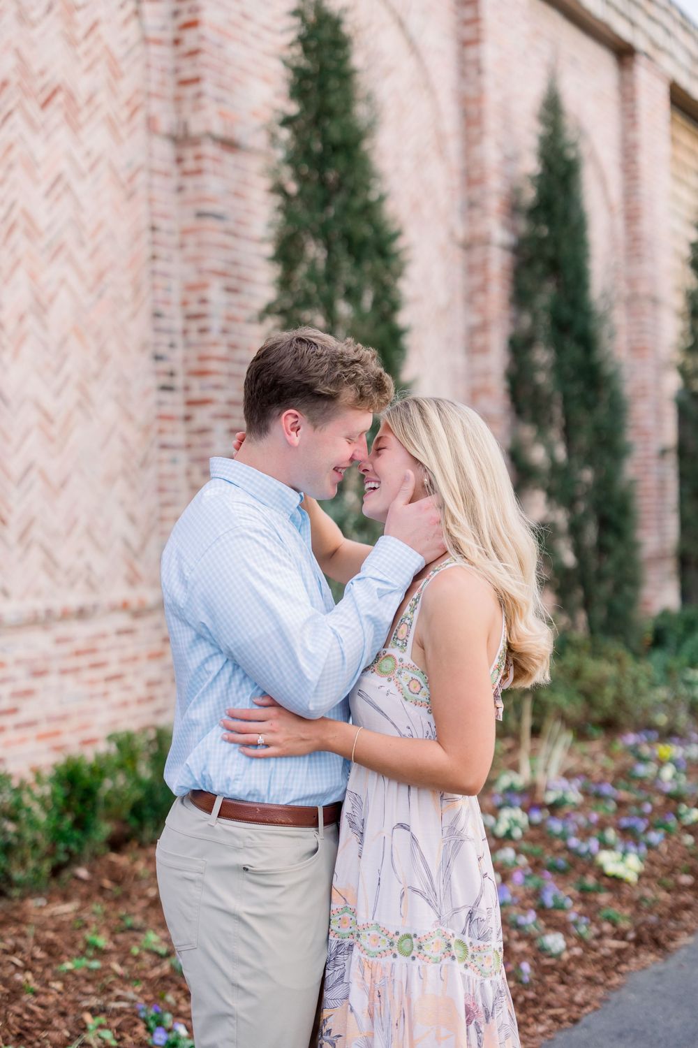 A romantic couple shares an intimate moment against a brick wall with evergreen trees and flowers in the background.