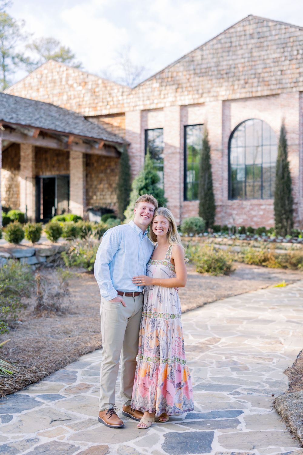 A couple shares a romantic moment on a stone patio outside a rustic brick building in warm afternoon light.