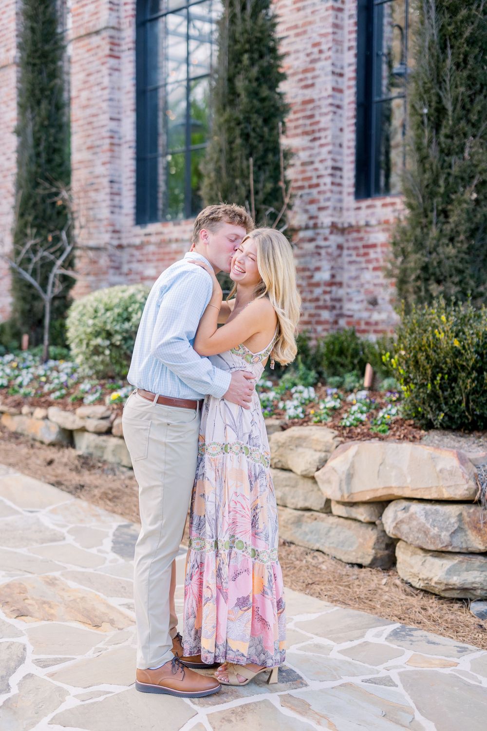 Romantic couple sharing tender moments against a rustic brick wall backdrop with landscaped garden.