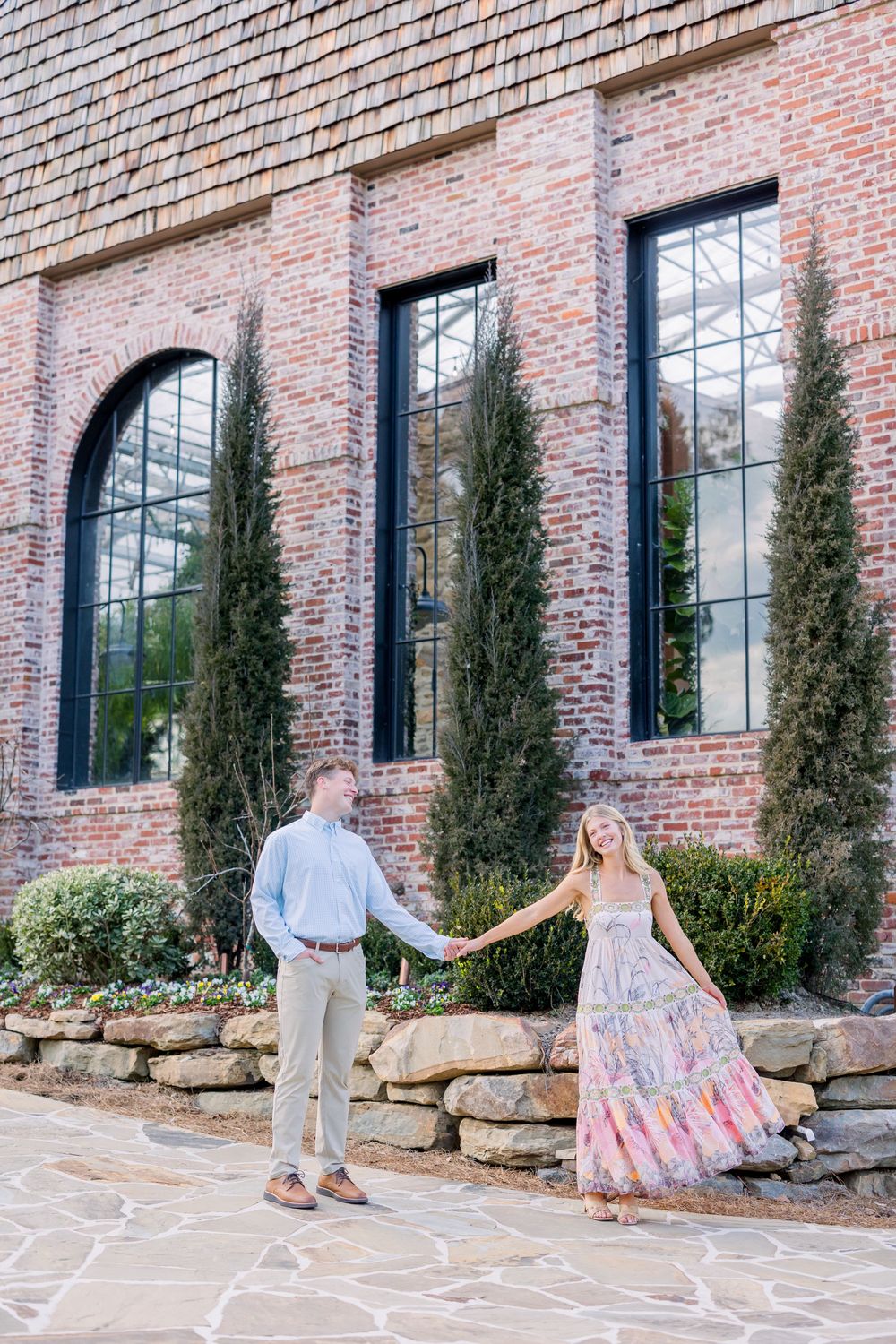 Couple dances playfully in front of a rustic brick building with tall cypress trees and stone landscaping.