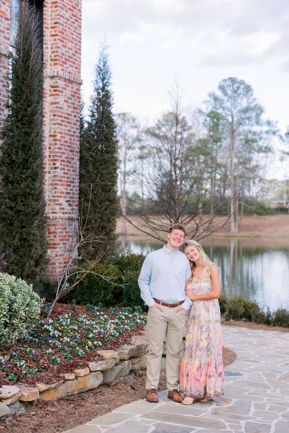 Couple poses together by a lakeside brick venue with landscaped gardens and cypress trees.