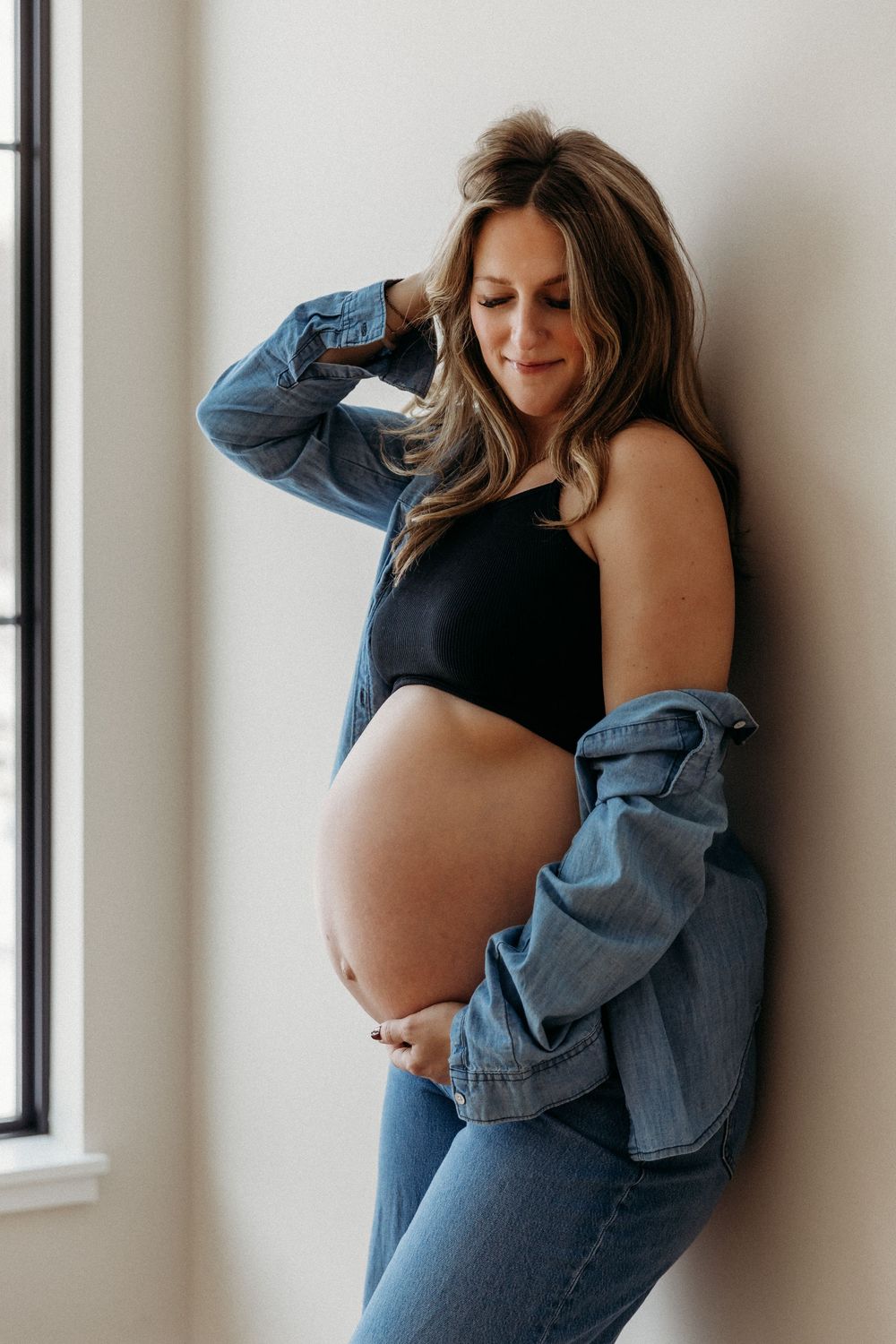 Window-lit maternity portraits with casual denim and black ensemble.