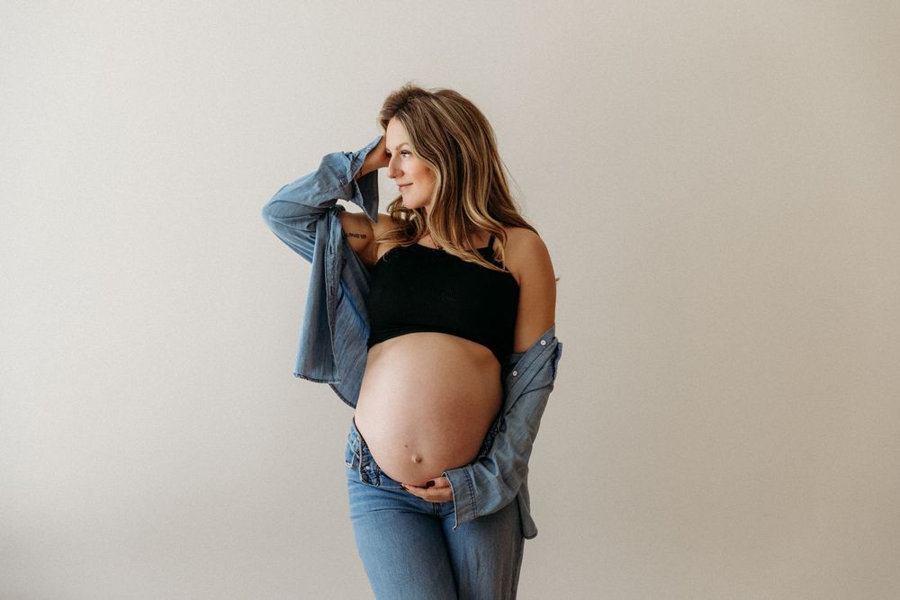 Maternity photo of a pregnant woman wearing denim and black against a light wall.