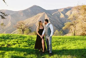 A couple shares romantic moments in a scenic mountain landscape with green grass and golden sunlight.
