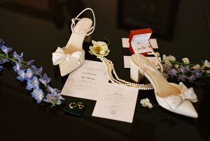Wedding accessories including white bow heels, invitation, jewelry and delicate blue flowers arranged on dark surface.