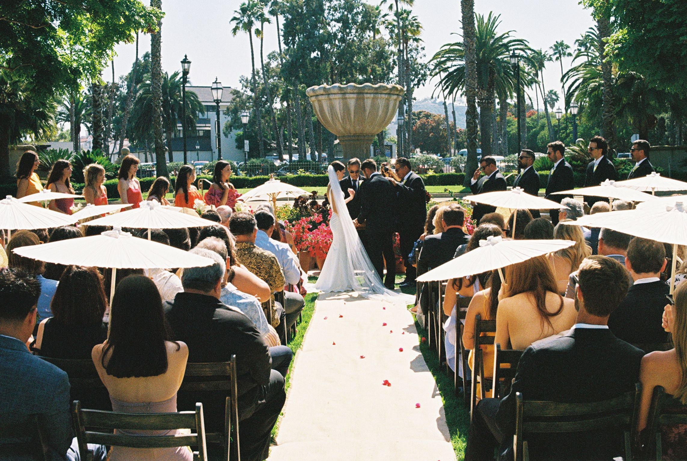 Rows of attendees seated outdoors under white umbrellas at a daytime event with palm trees in the background.