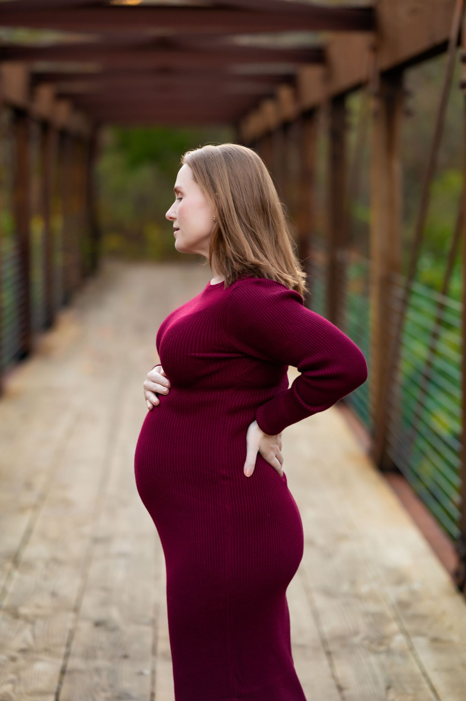burgundy dress compliments mom-to-be's silhouette during maternity photo session; mom-to-be standing on a rustic brown bridge