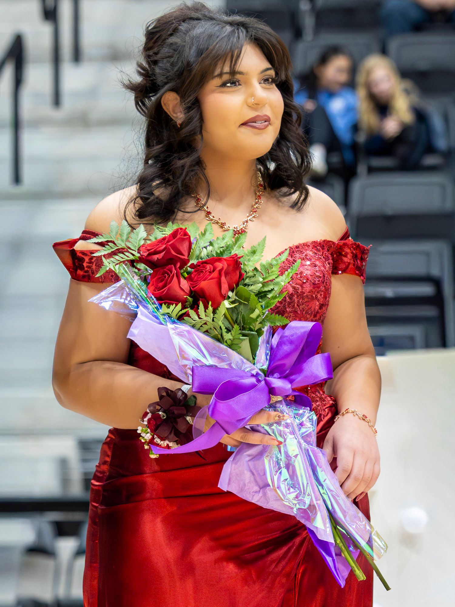A performer in a sparkling red formal dress holds a vibrant bouquet of red roses and purple flowers on stage.