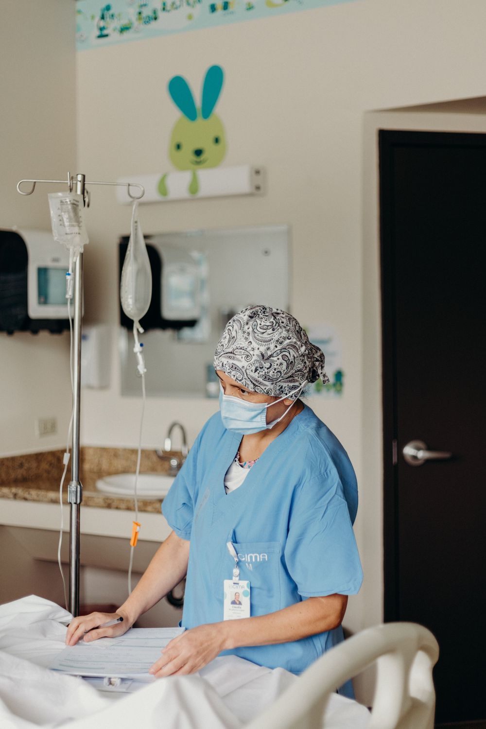 Healthcare worker in blue scrubs adjusts bedding and medical equipment in a hospital room.