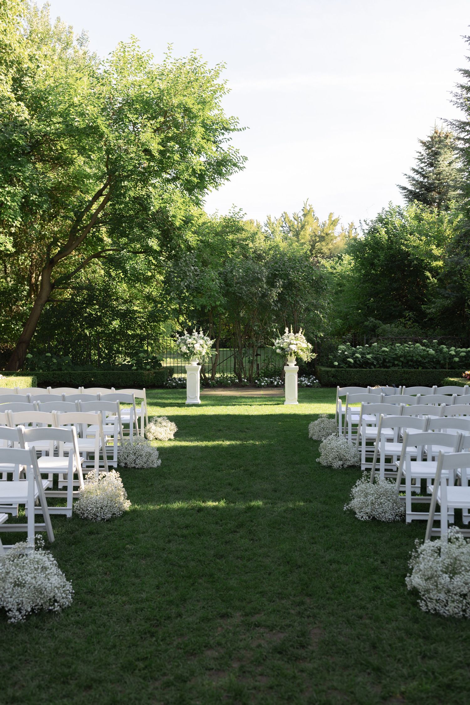 White chairs and floral arrangements line a grassy outdoor wedding ceremony space under green trees on a sunny day.