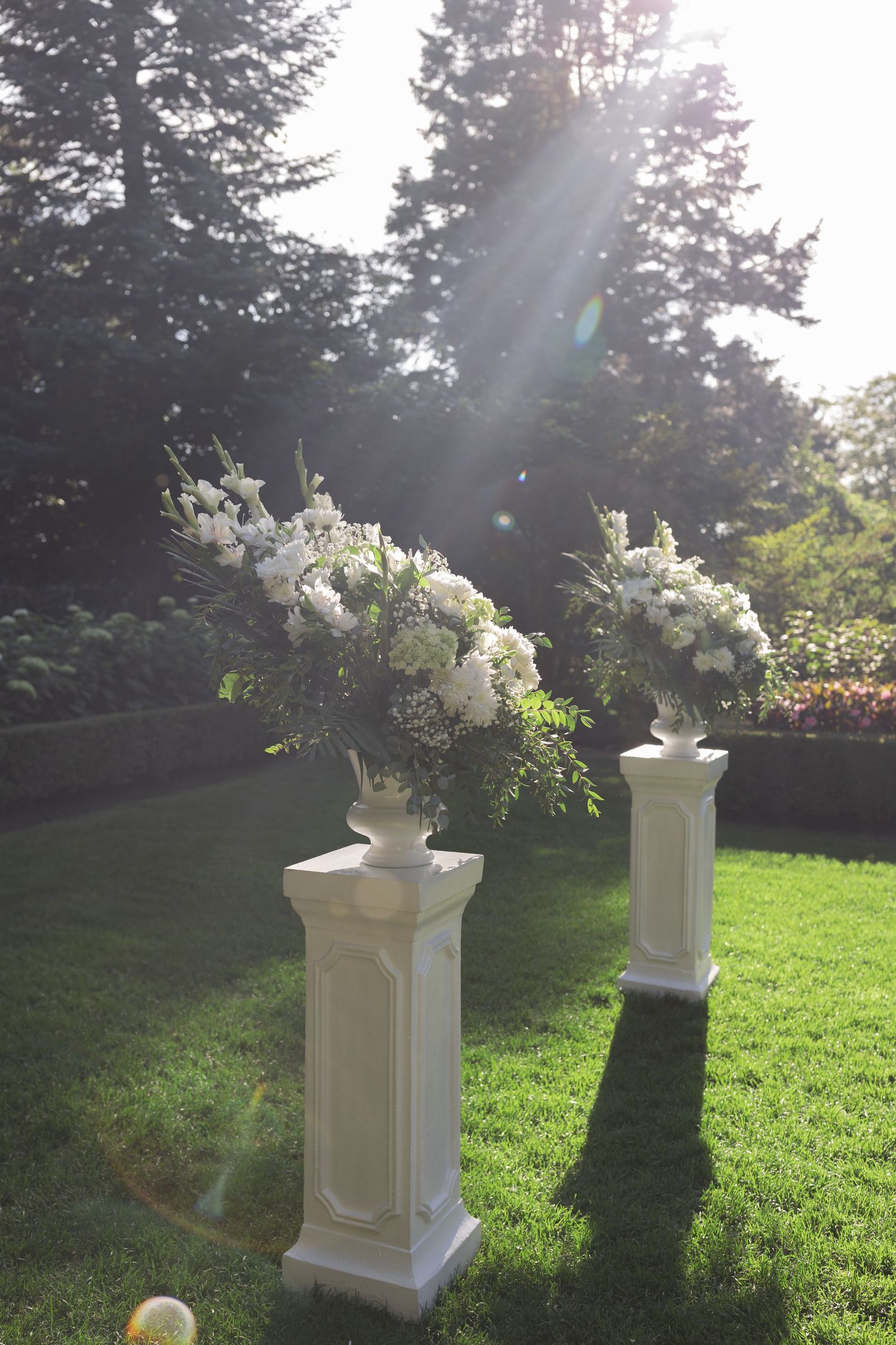 White decorative pedestals with floral arrangements line a garden path in dappled sunlight.