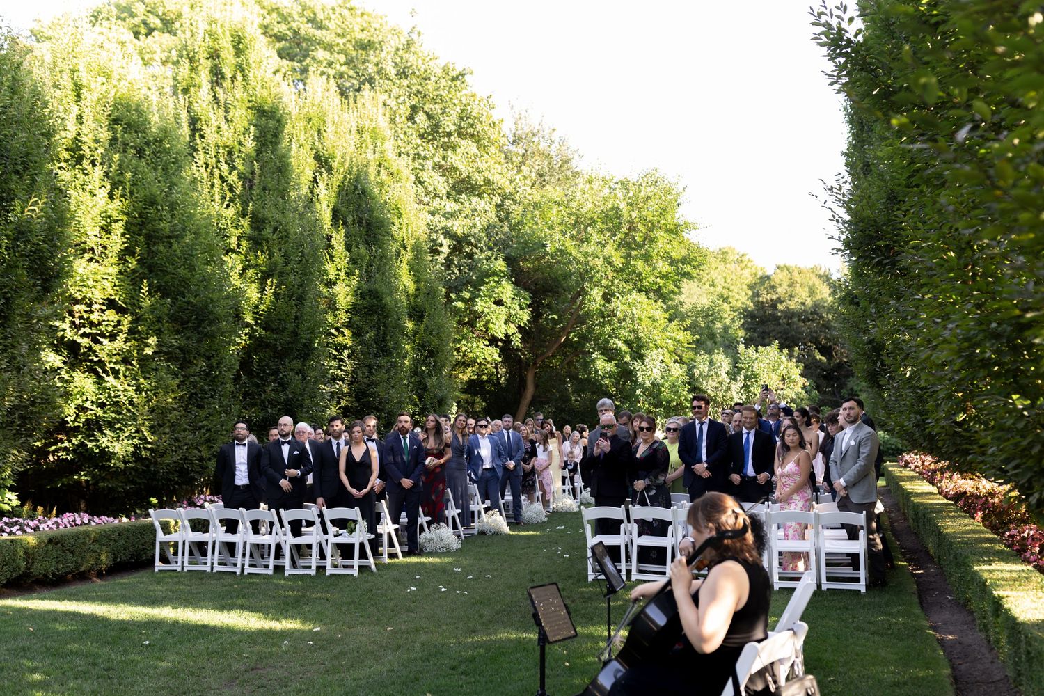 Guests seated in white chairs for an outdoor garden wedding ceremony beneath towering willow trees.