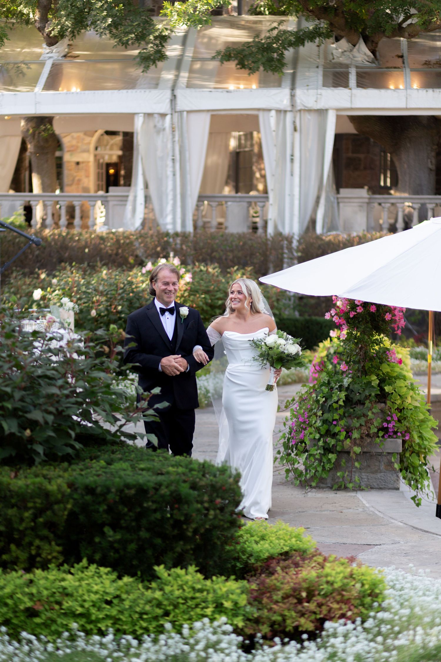 A couple walks through a garden pathway with white drapes and floral arrangements at an elegant outdoor wedding venue.