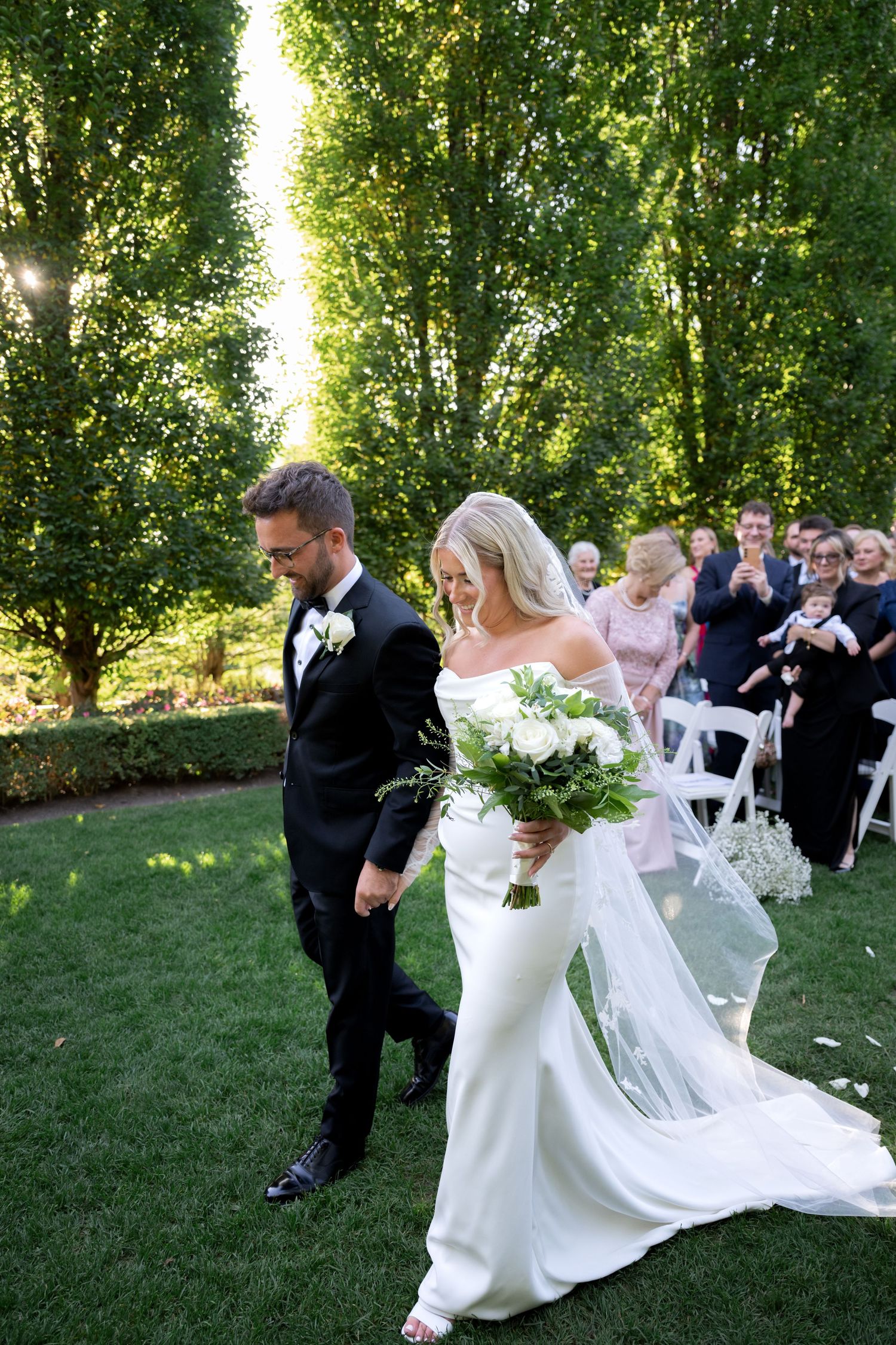 A couple dances playfully on a lush green lawn surrounded by wedding guests and tall trees.