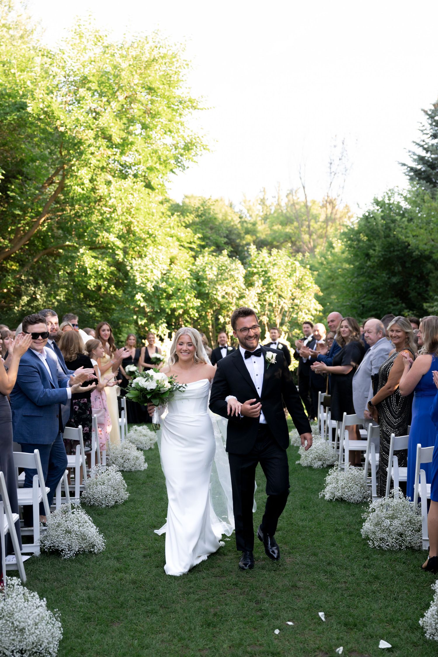 Wedding guests celebrate outdoors at a garden ceremony with white chairs and baby's breath floral decorations.