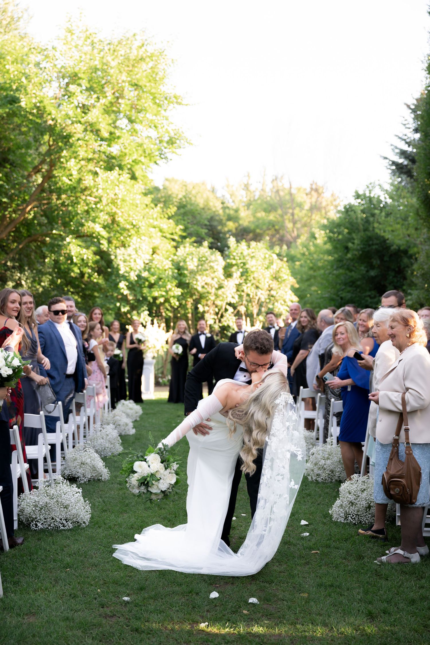 Wedding guests line up outdoors to watch a couple's celebratory dance on a sunny garden lawn.