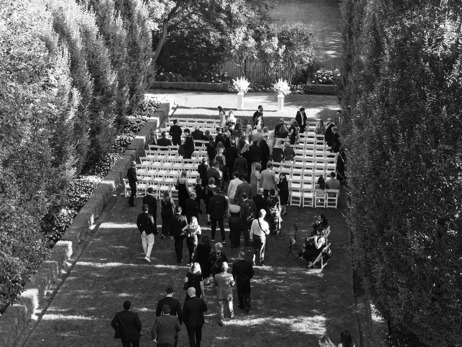 People walk along a tree-lined path in a park with sunlight filtering through the branches.