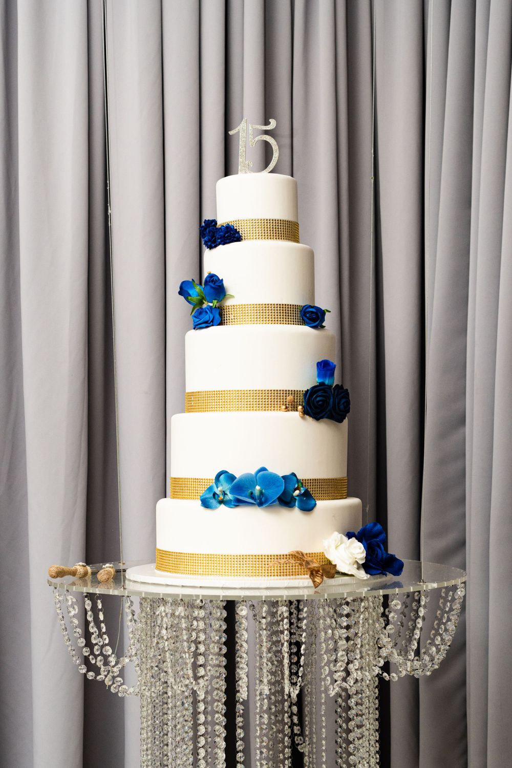 Three-tiered white wedding cake with gold bands and blue floral accents on a crystal cake stand.