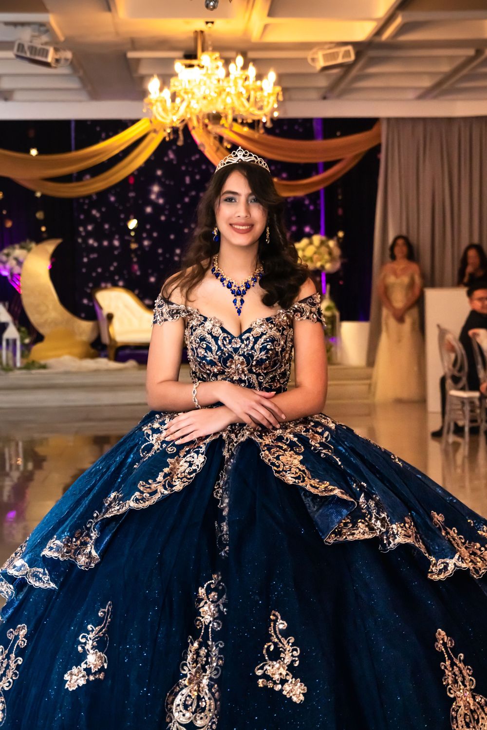 A stunning navy blue and gold quinceañera ball gown under an elegant chandelier.