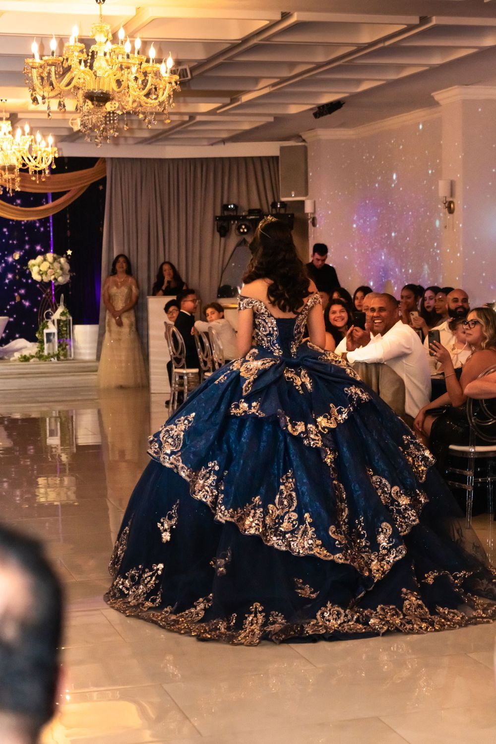 Ornate navy blue and gold gowns swirling on a white dance floor under crystal chandeliers.
