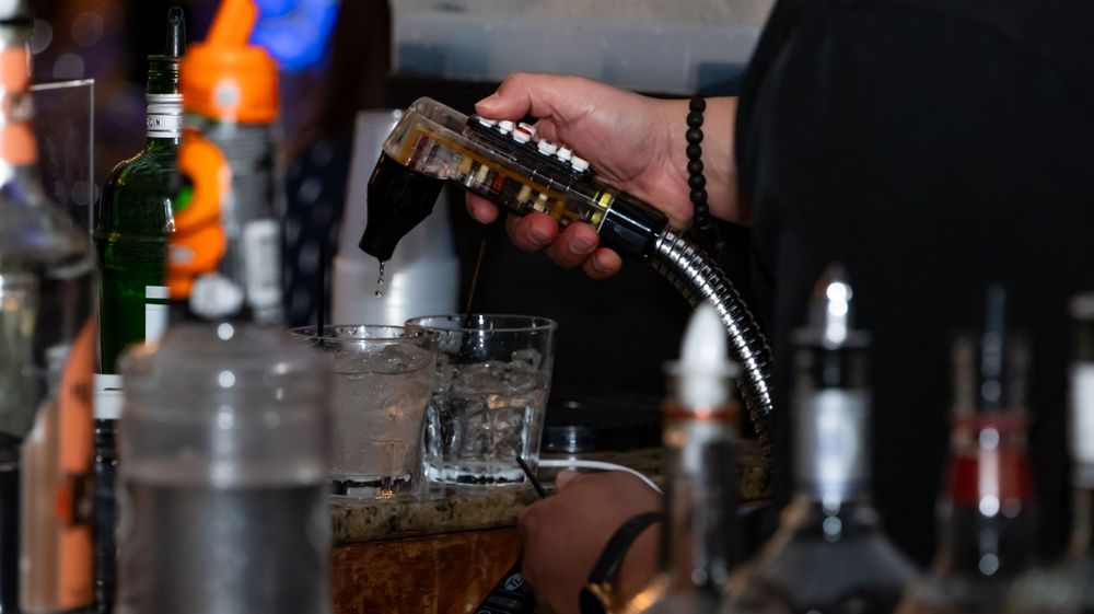 Bartender pouring a drink at a bar with bottles in the background.