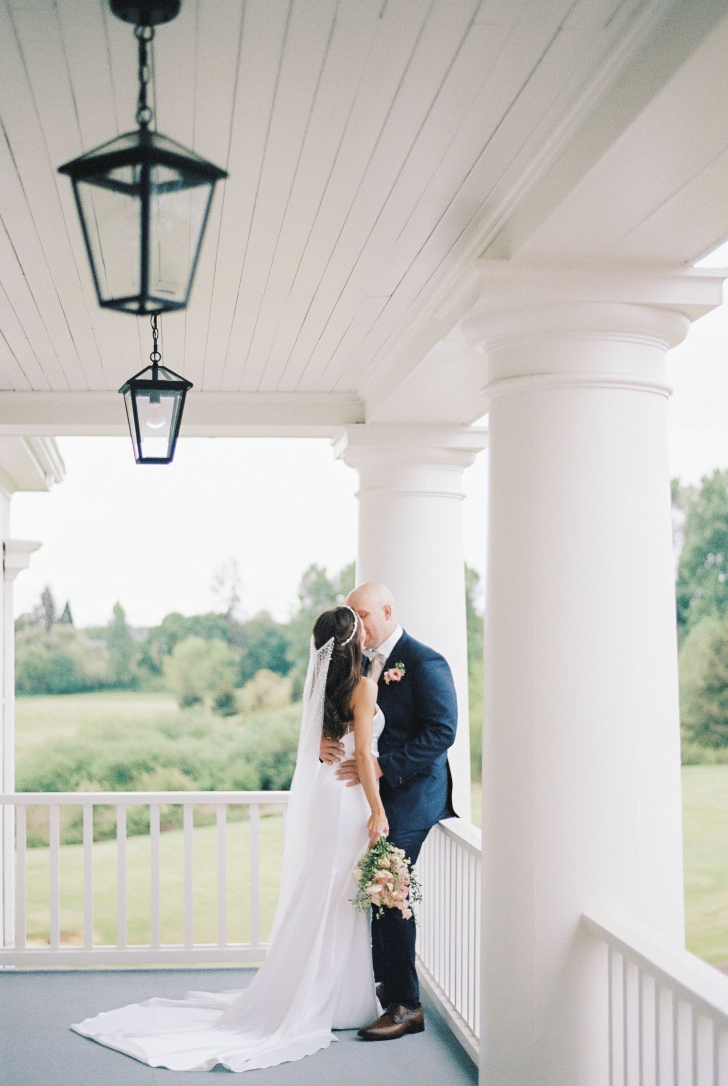 Wedding couple embraces on a white columned porch with hanging lanterns and scenic countryside views.